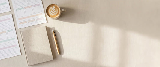 Cozy flat lay of pastel printable worksheets, a beige notebook, gold pen, and a latte on a soft neutral desk with warm sunlight.