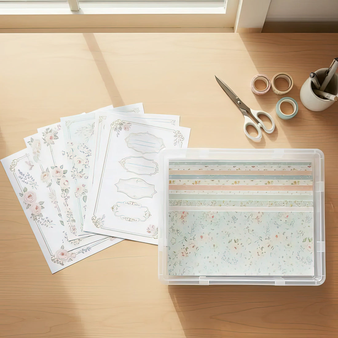 Neatly organized printed clipart sheets and digital papers stored in a clear scrapbook box on a tidy craft desk, with scissors and washi tape beside them.
