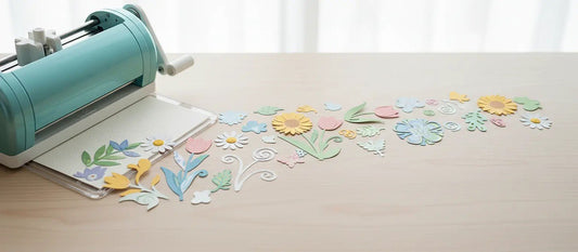 Manual die-cutting machine on a desk with colorful paper flowers and leaf die-cuts scattered in a row.