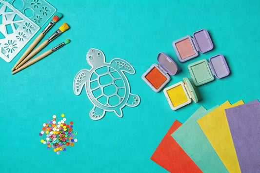 Flat lay on a teal background with a turtle stencil, paintbrushes, small ink pads, colorful cardstock sheets, and a pile of sequins.