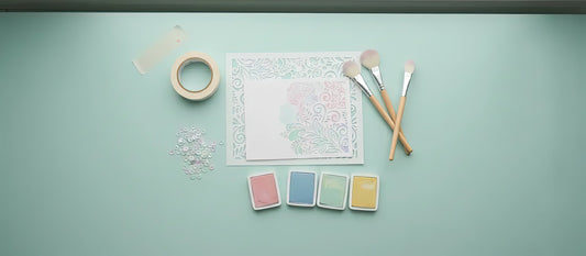 Flat lay of stencil tools with blending brushes, ink pads, and a stenciled card panel on a pastel teal background.
