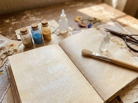 Cozy junk journal spread on a wooden desk with shimmery blank pages, small glass jars of blue and gold mica powder, a paintbrush, and tiny glue bottles ready for crafting.