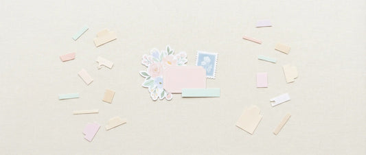 Flat lay of a cozy crafting desk with a small layered paper cluster made from floral clipart, a label, a word strip, and faux postage, surrounded by neatly scattered pastel paper pieces on a soft neutral background.