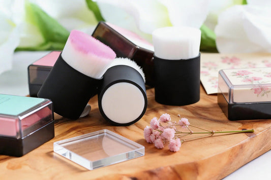 Ink blending brushes and pastel ink pads on a wooden craft board with a clear acrylic stamping block and small pink flowers.