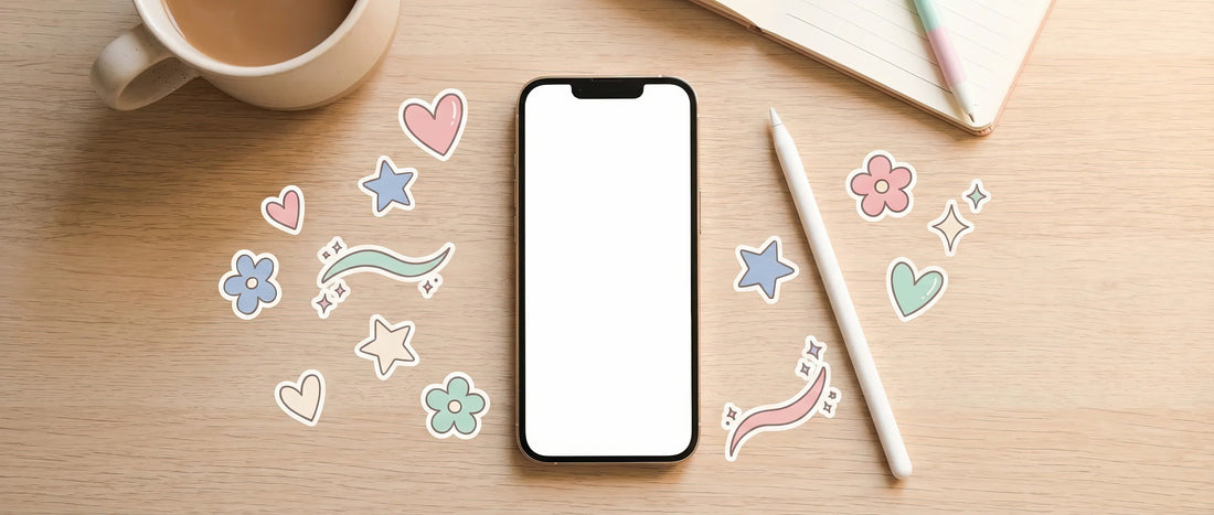 Top-down view of a smartphone with a blank white screen on a wooden desk, surrounded by pastel clipart stickers (hearts, stars, flowers and swooshes), a white stylus, a notebook and a coffee cup.